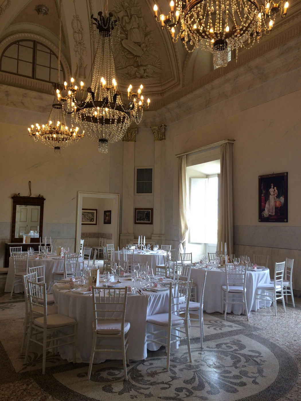 A grand marble ballroom under crystal chandeliers, round tables set in white for the wedding