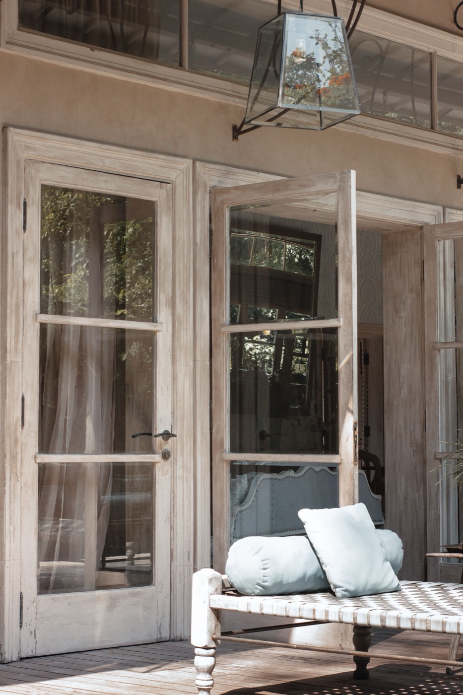 Weathered French doors open onto a tropical deck, sheer curtains and a daybed beyond