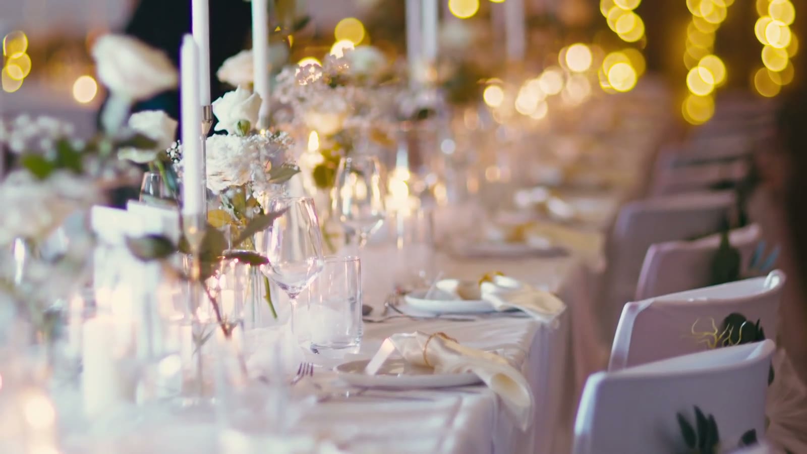 A long candlelit table set for a wedding — taper candles, white flowers, and warm string lights beyond