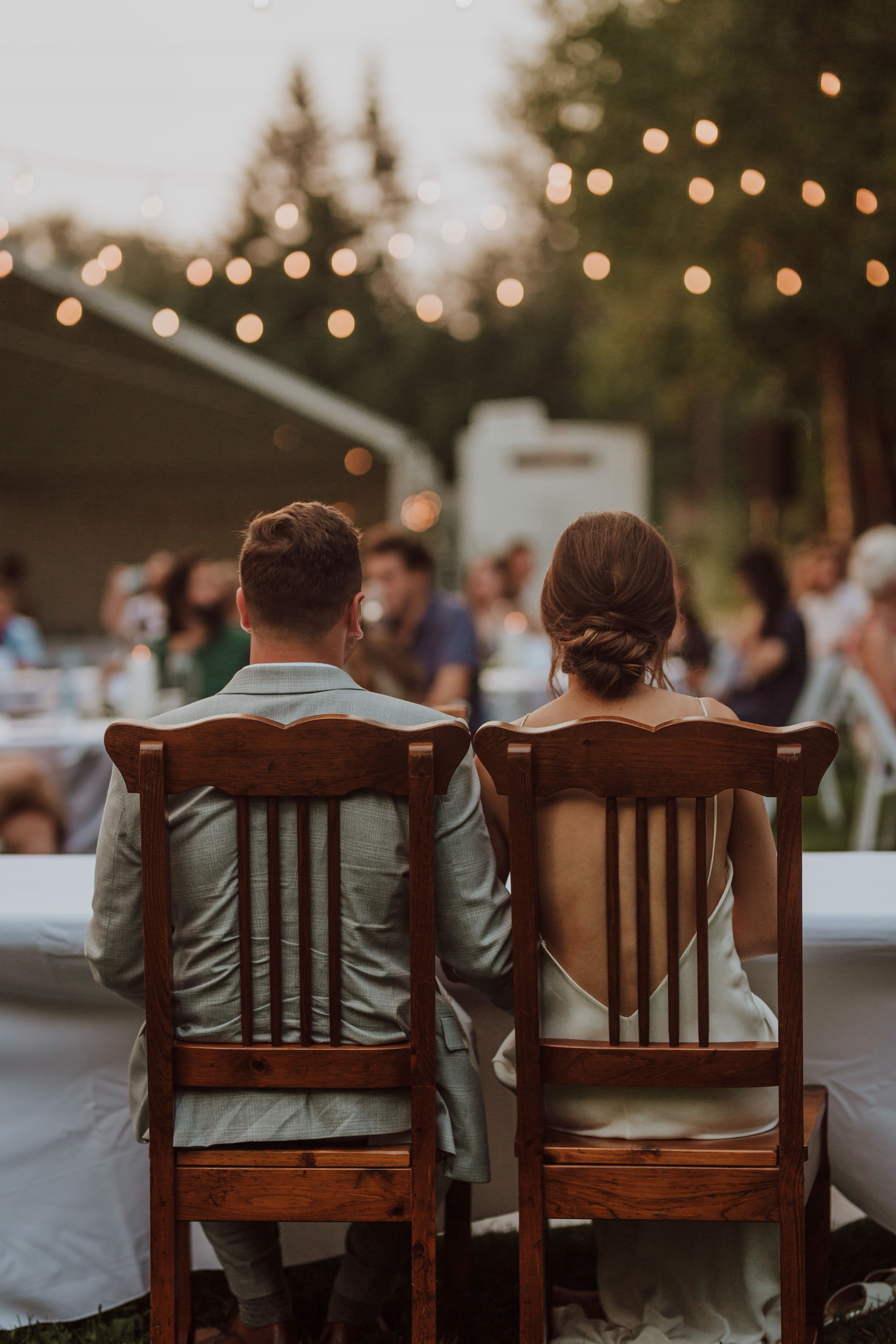 A couple seated together at a sweetheart table at dusk, backs to the camera, string lights overhead and guests at long tables beyond them