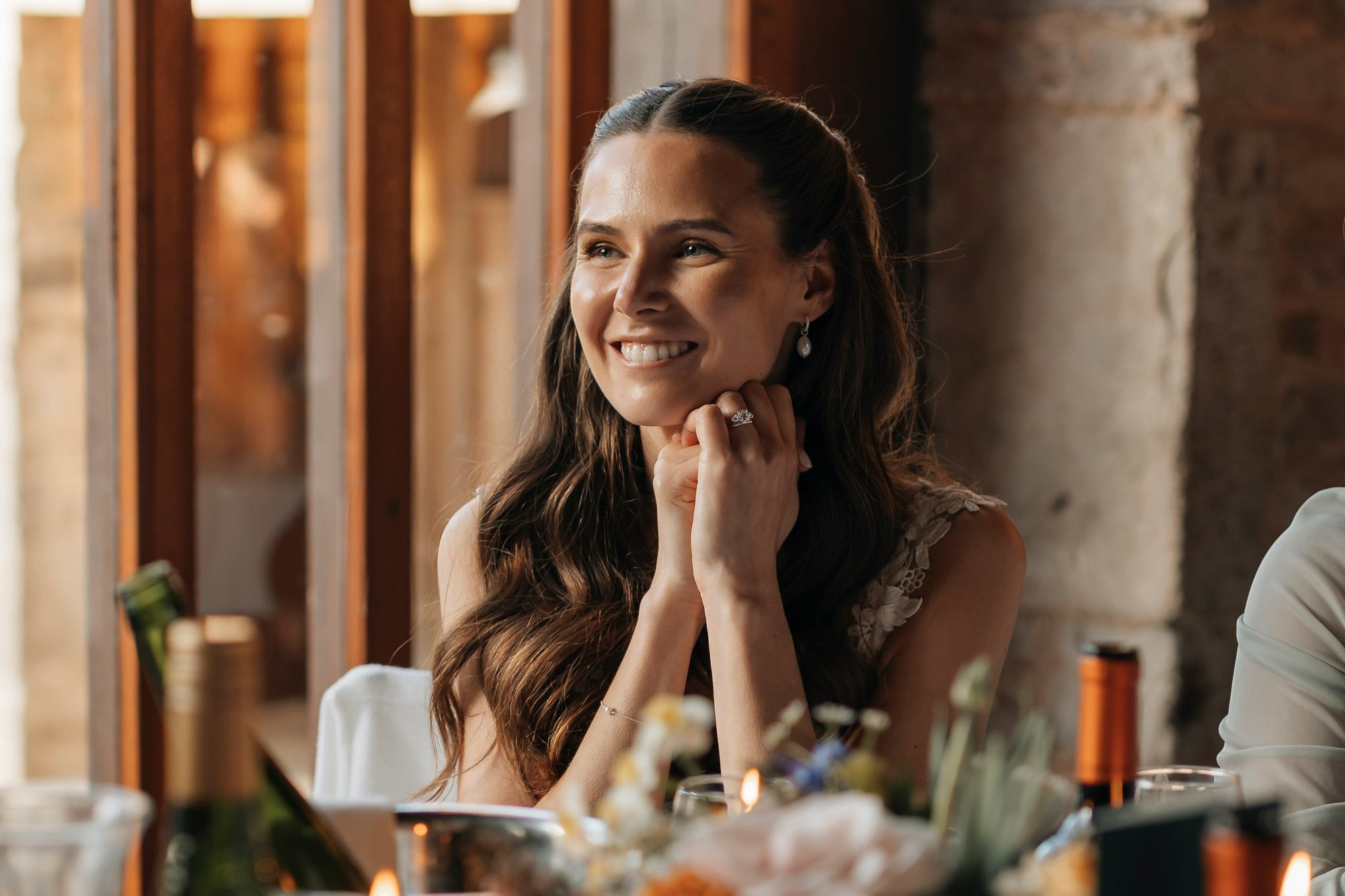 A bride at a candlelit wedding dinner table, chin resting on her hands, laughing at something outside the frame.