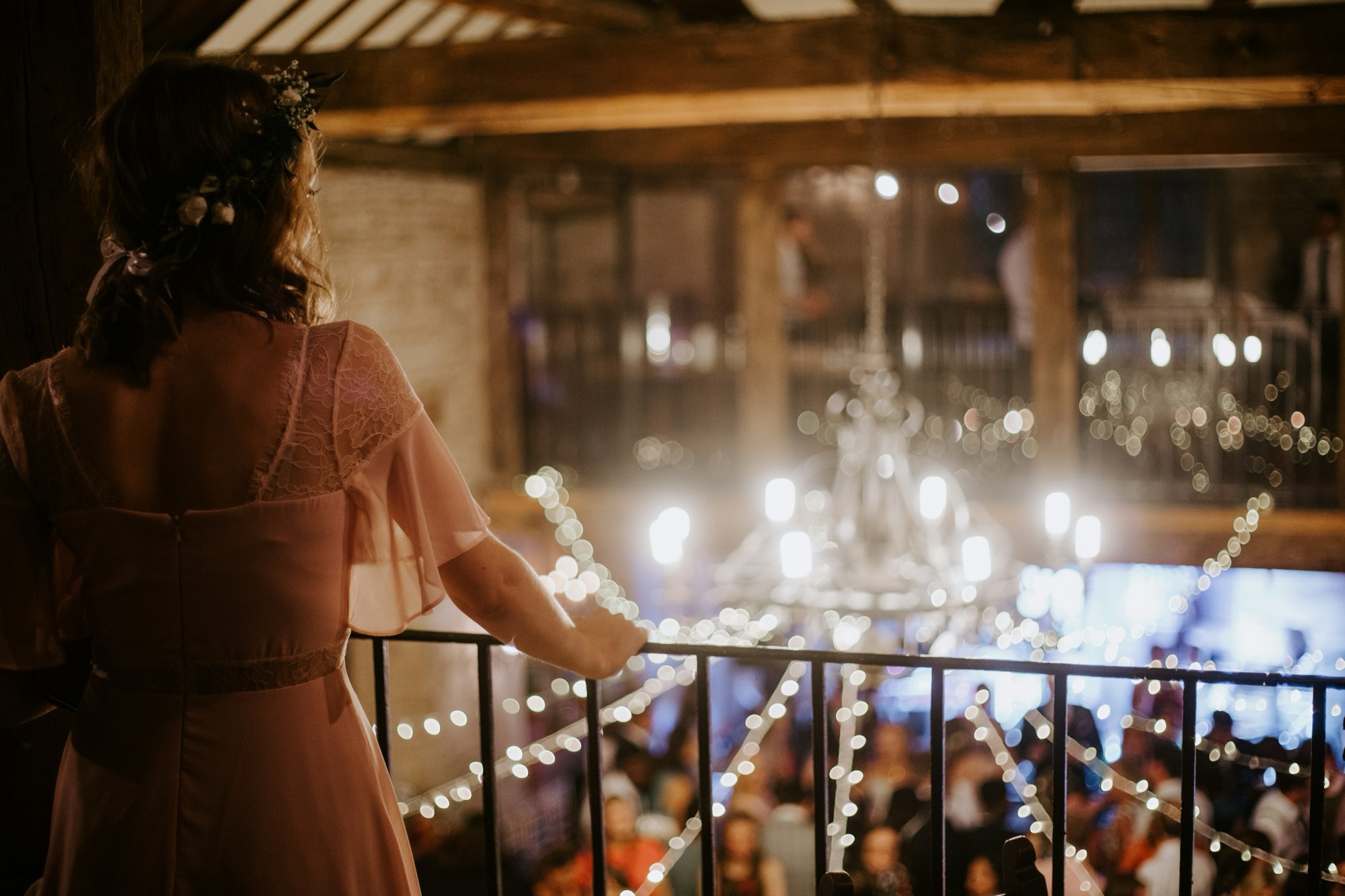 A guest in a blush dress stands at a mezzanine rail, seen from behind, looking down over a warmly lit reception of fairy lights and a chandelier
