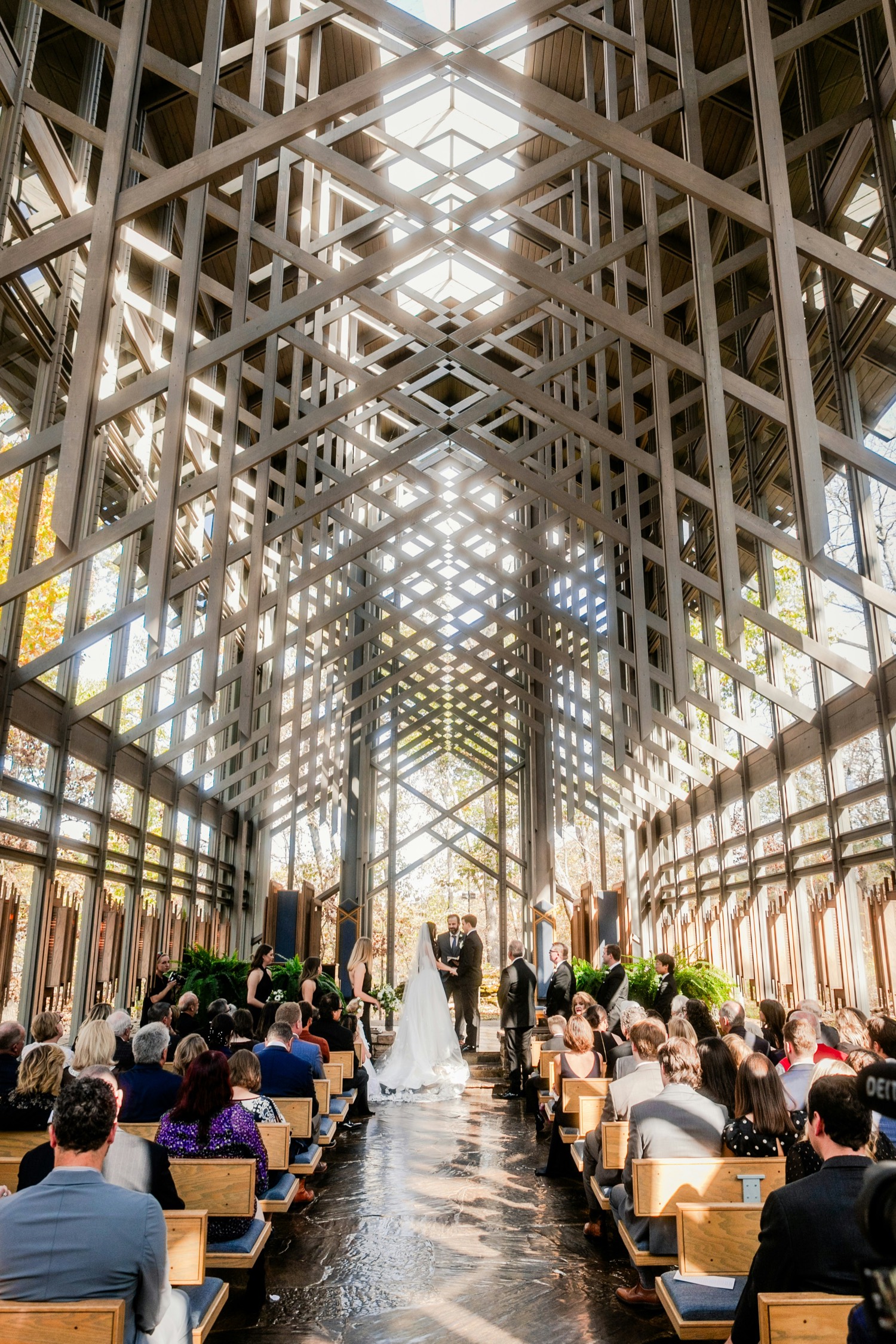 A congregation seated in a glass-and-timber chapel, witnessing a wedding ceremony down a sunlit aisle